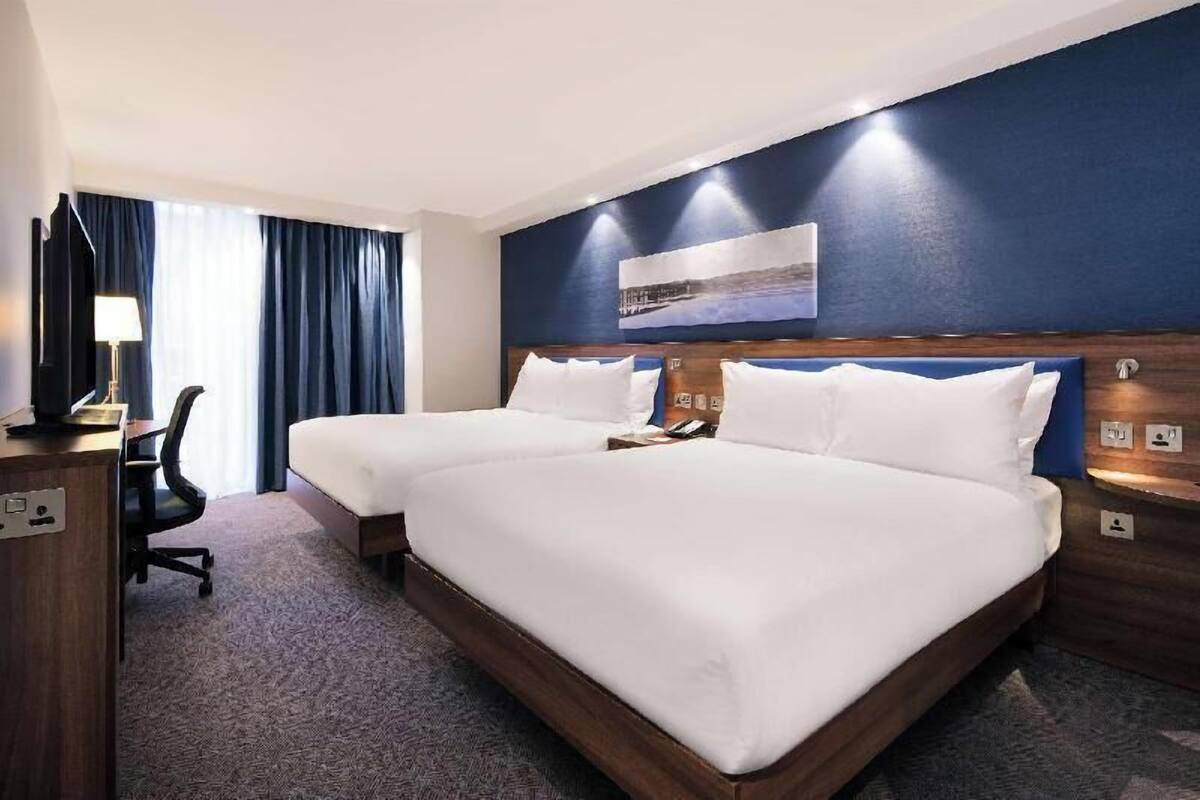 A hotel room with two beds, a desk, a chair, and a large window with curtains.