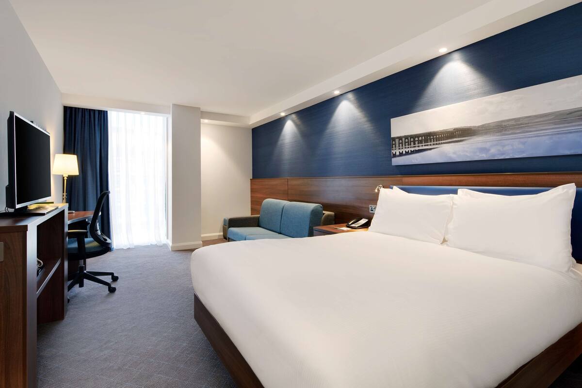 A hotel room with a large bed, a desk with a computer, a chair, a TV, and a painting on the wall.