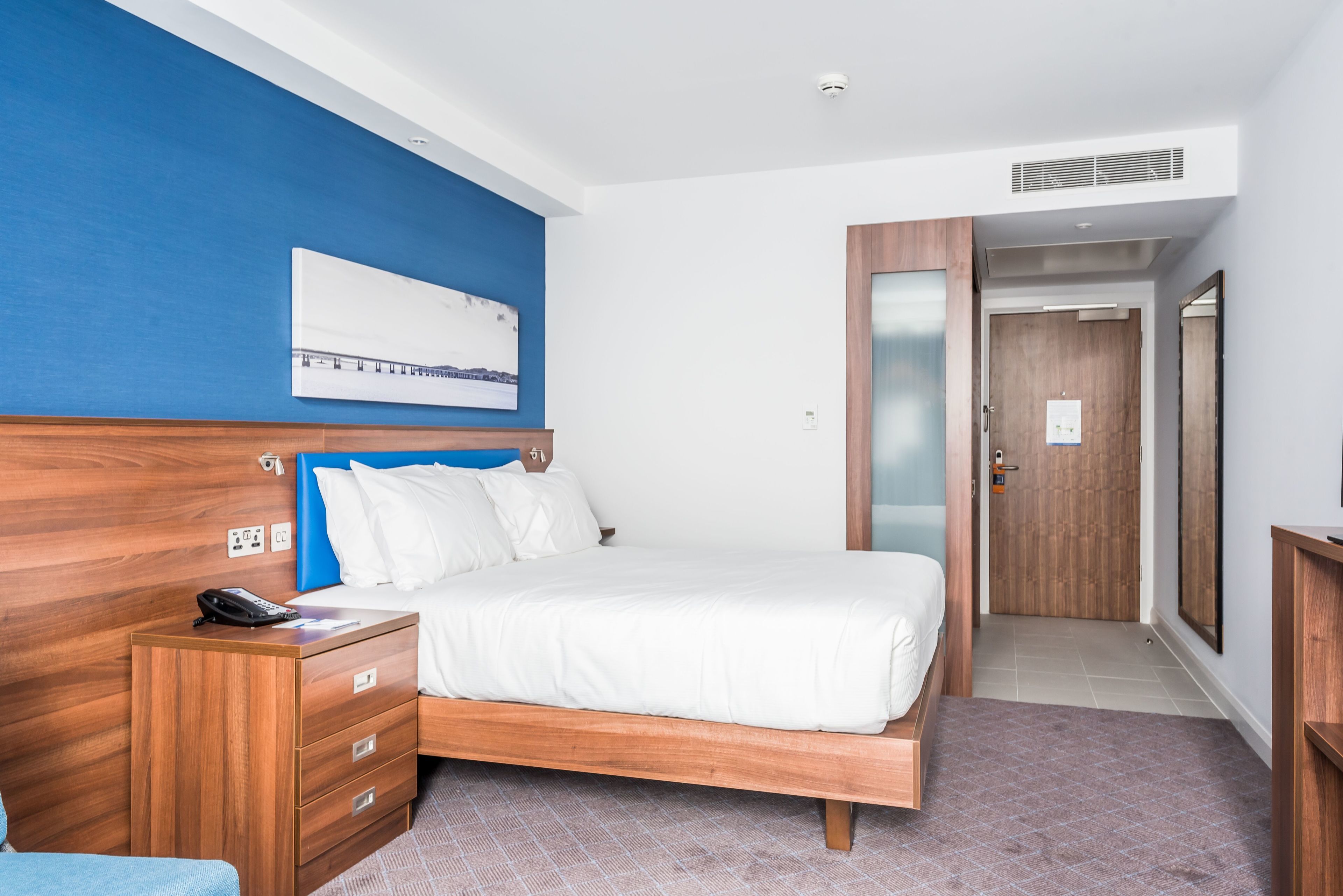 A hotel room with a wooden bed, a blue accent wall, a white headboard, a bedside table with a phone, and a door leading to a hallway.