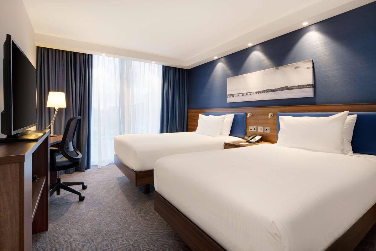 A hotel room with two beds, a desk with a chair, a TV, and a painting on the wall.