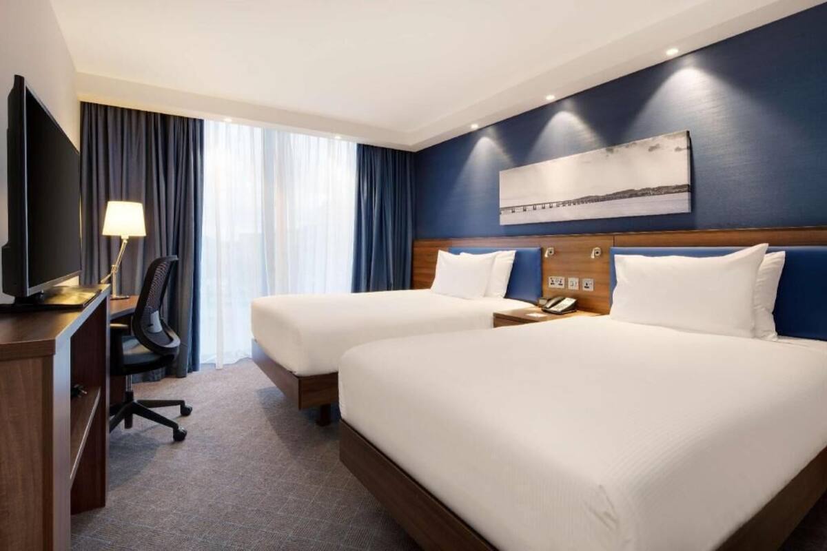 A hotel room with two beds, a desk with a chair, a TV, and a painting on the wall.
