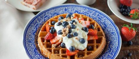 Un plato de waffles cubiertos de fresas, arándanos y crema batida, acompañado de una taza de café y una porción de pastel sobre una mesa de madera.
