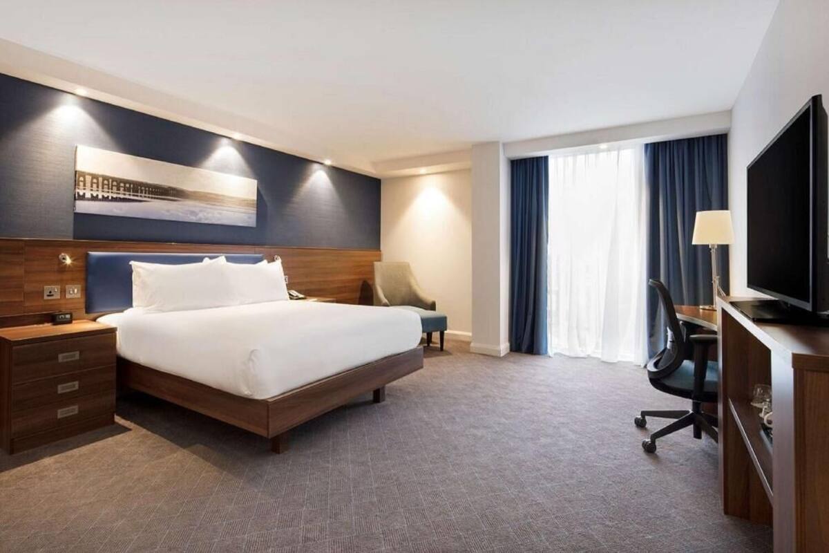 A hotel room with a large bed, a desk with a TV, a chair, and a window with curtains.