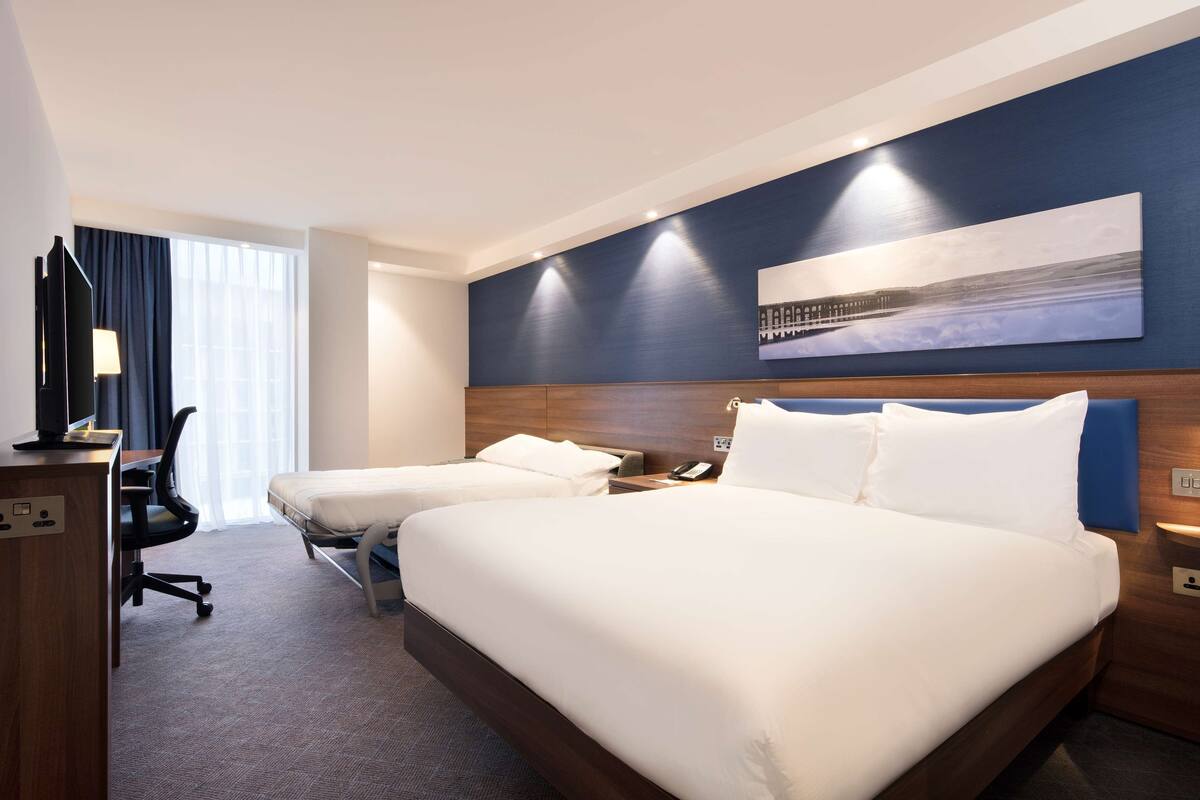 A hotel room with two beds, a desk, a chair, and a large window with curtains.