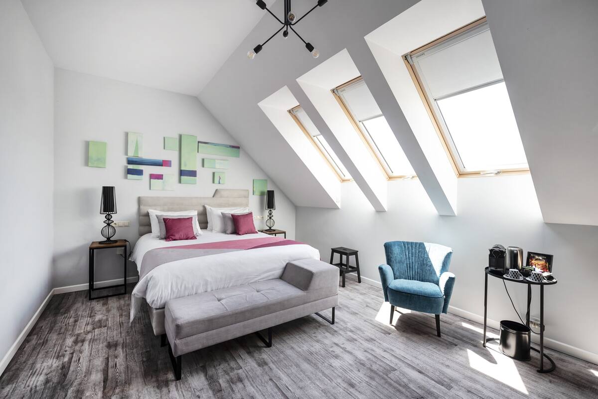Ein modernes Schlafzimmer mit einem großen Bett, einem blauen Sessel, einem kleinen Tisch mit Wasserkocher und Dachfenstern.