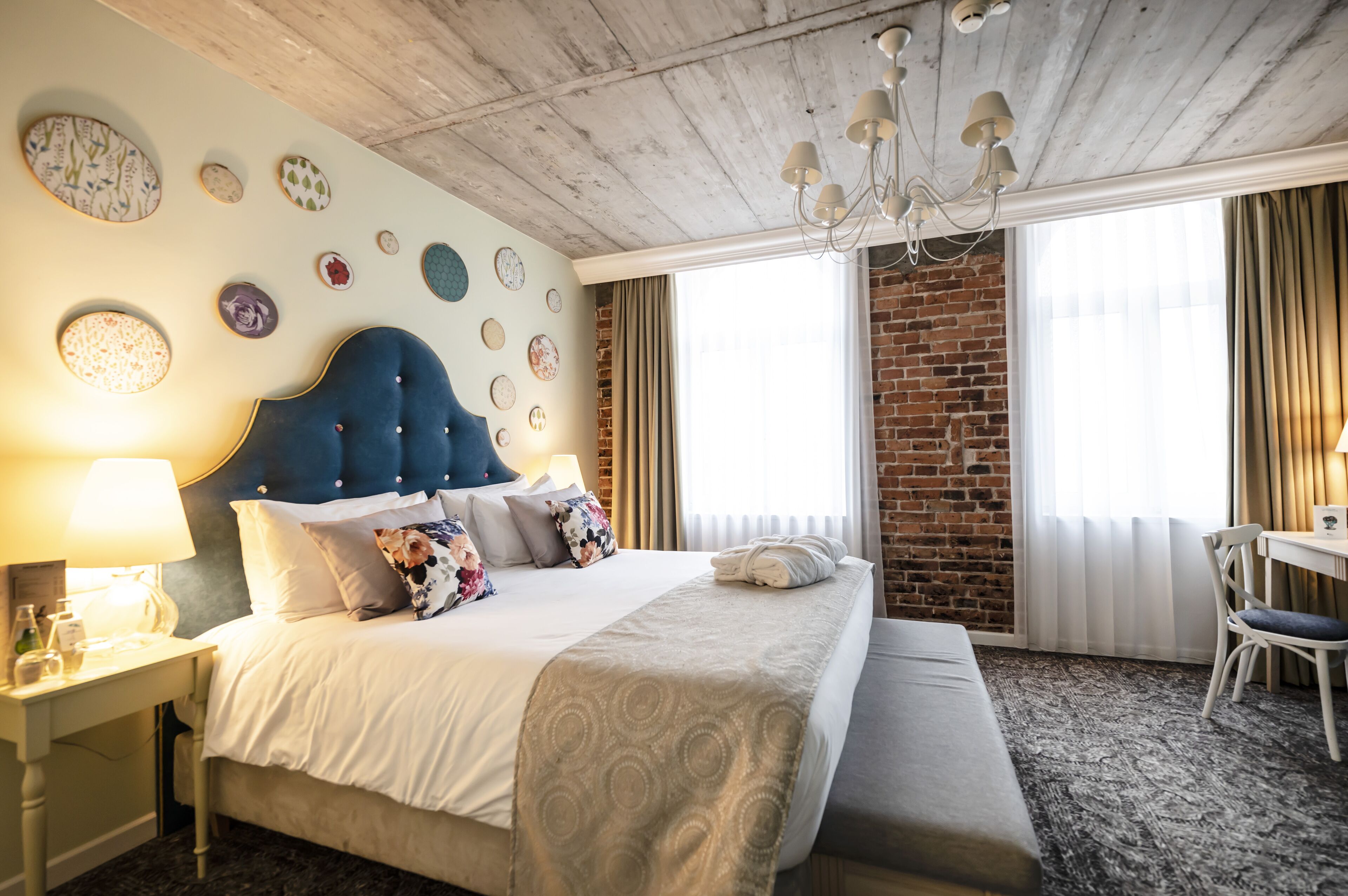 Quarto de hotel com uma cama grande, uma escrivaninha e uma cadeira. O quarto possui uma parede de tijolos e um lustre.