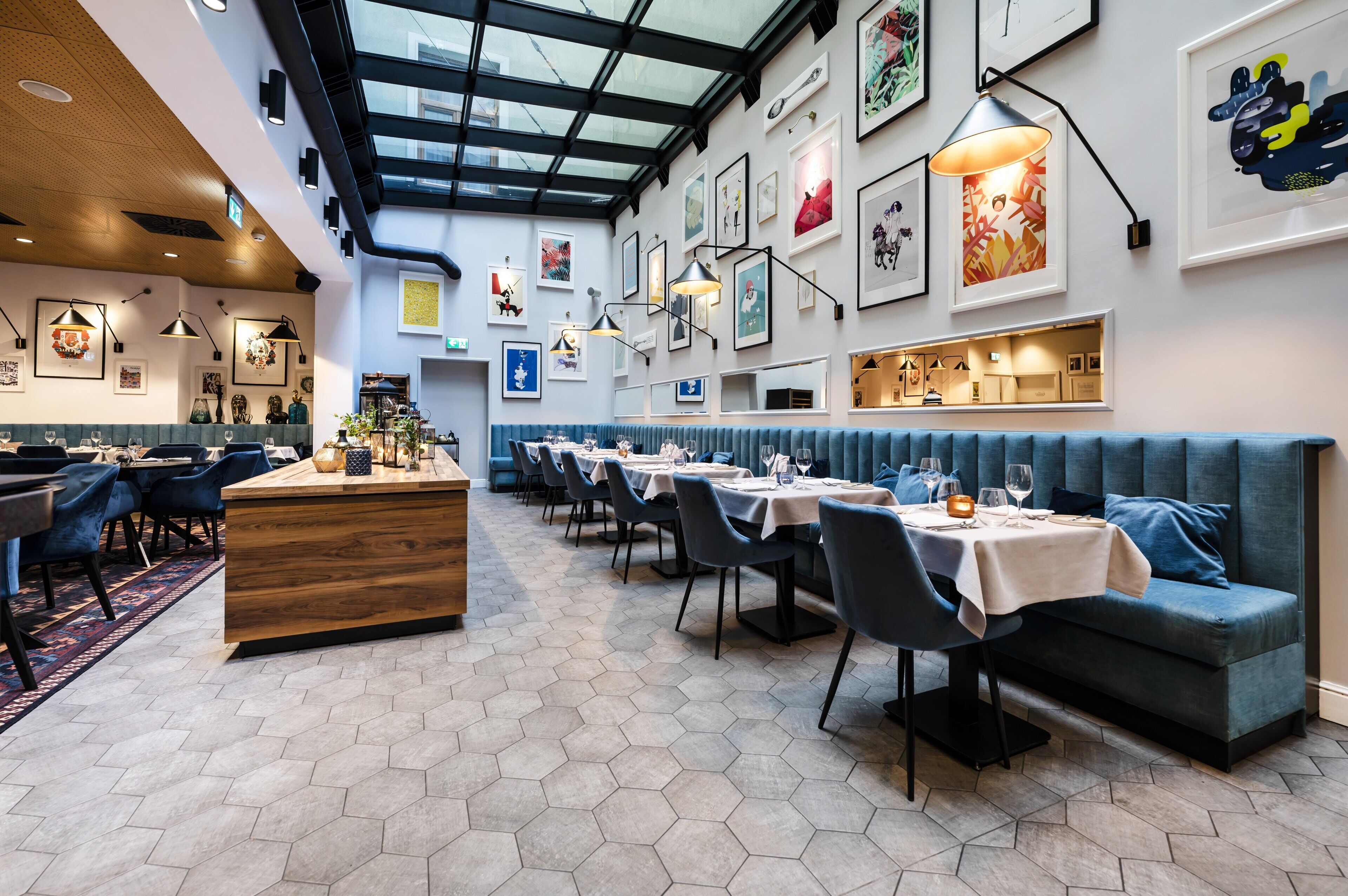 A restaurant with hexagonal tile flooring, a central wooden counter, blue upholstered booths, and a variety of framed artwork on the walls.