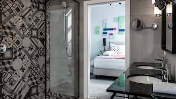 A modern bathroom with a glass shower enclosure, a black marble sink, and a bed visible through an open door.