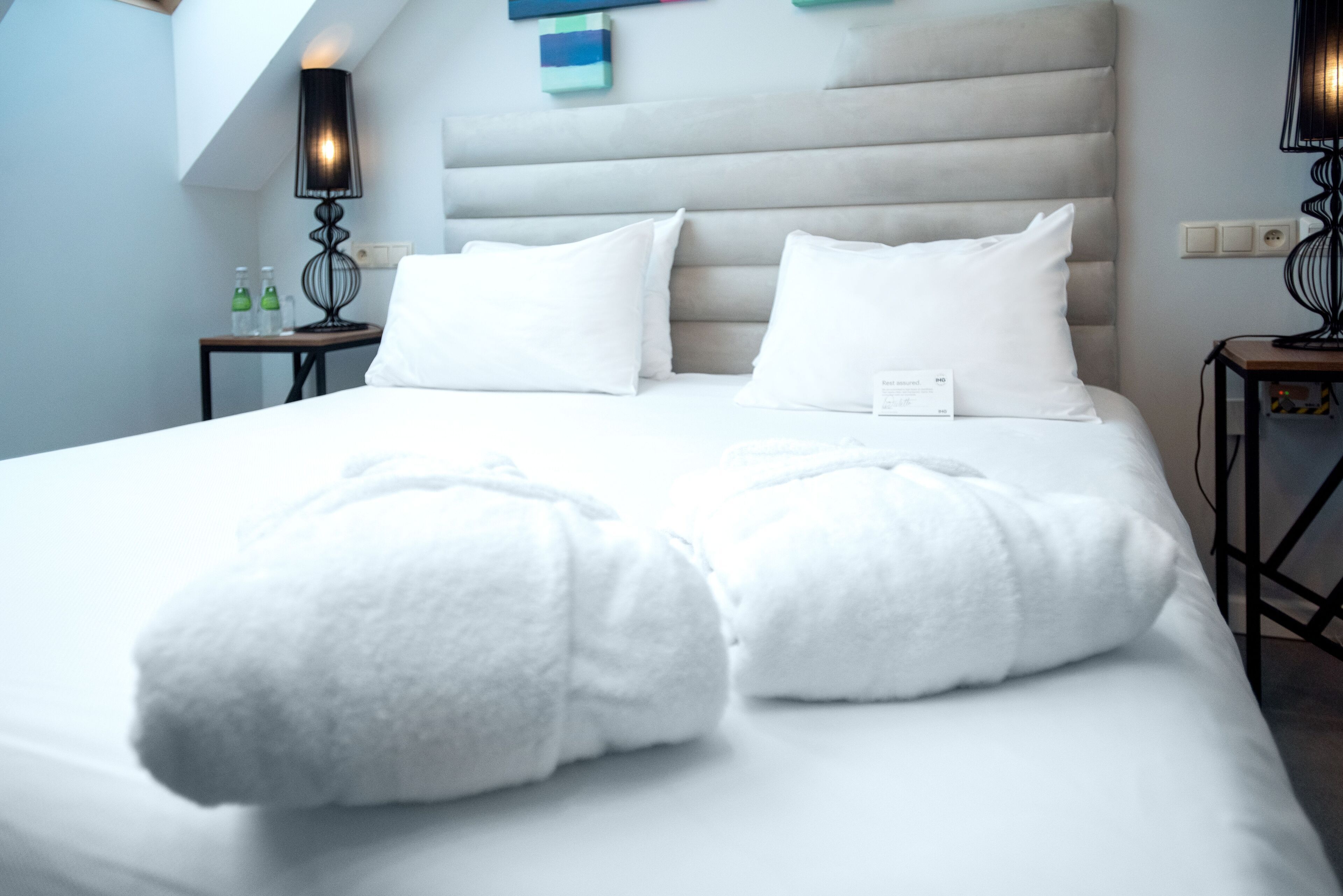 Cama bem arrumada com lençóis e almofadas brancas, cabeceira com painéis horizontais e duas mesas de cabeceira com abajures.