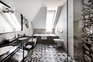 Une salle de bain moderne avec deux lavabos, une baignoire et des toilettes. Le sol et les murs sont recouverts de carreaux aux motifs géométriques, et une lucarne se trouve au-dessus de la baignoire.