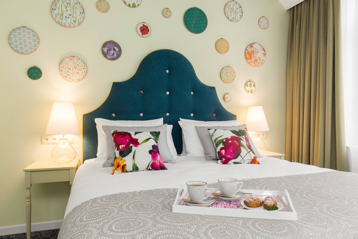 Ein ordentlich bezogenes Bett mit blumigen Kissen, ein Tablett mit Gebäck und Kaffee sowie eine Wand mit runden Kunstwerken.