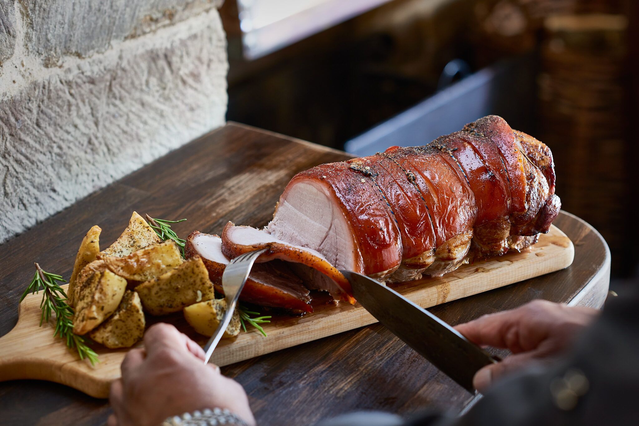 Un jambon rôti sur une planche à découper en bois, avec un couteau qui le tranche, entouré de pommes de terre rôties et de romarin.