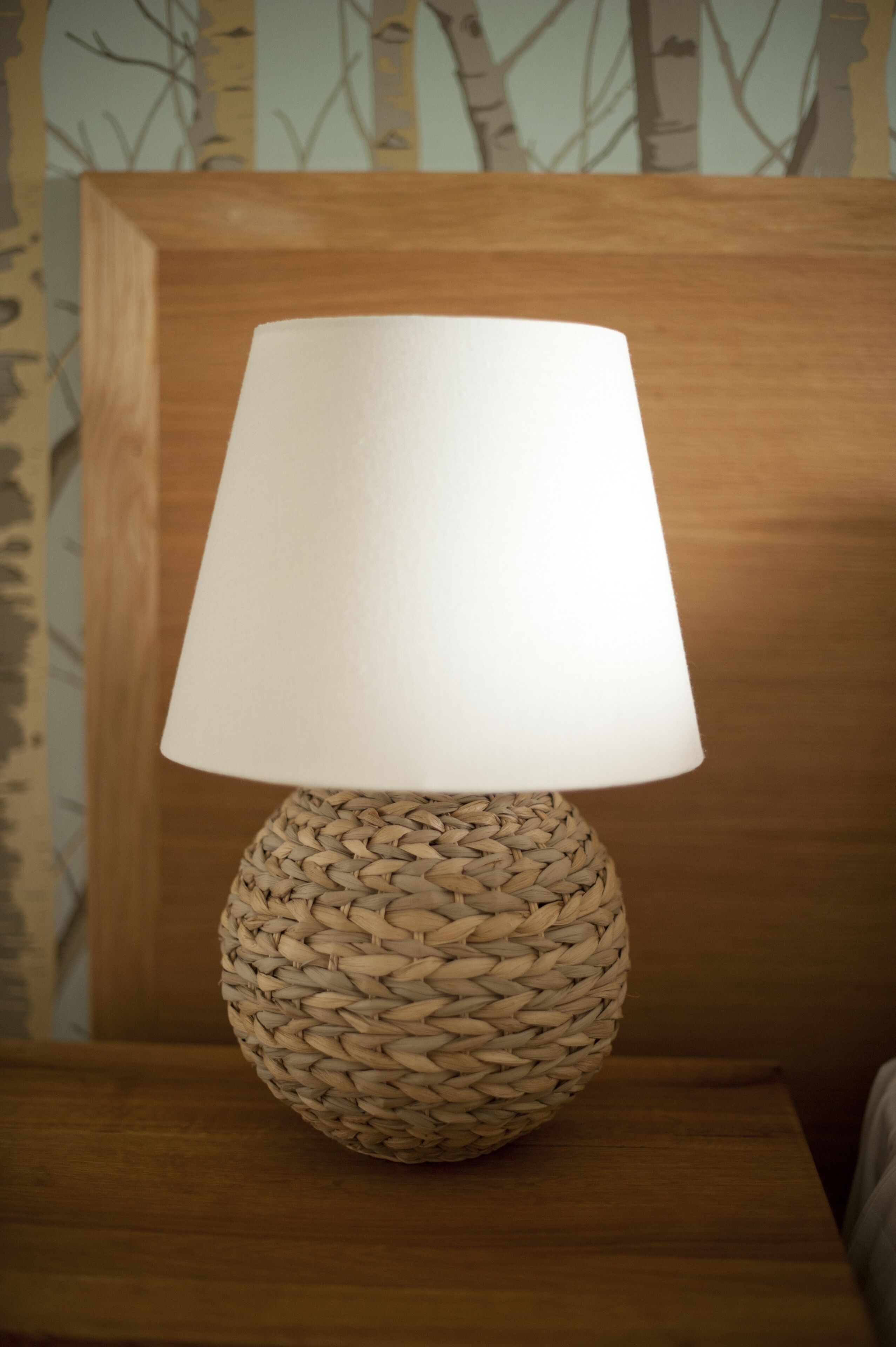 Una lámpara redonda trenzada con pantalla blanca sobre una mesita de noche de madera.
