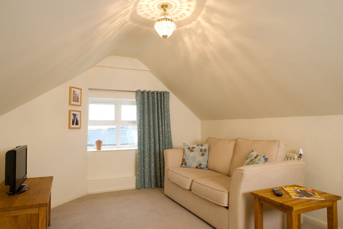 A cozy attic room with a beige sofa, a wooden table, a television, and a window with curtains.