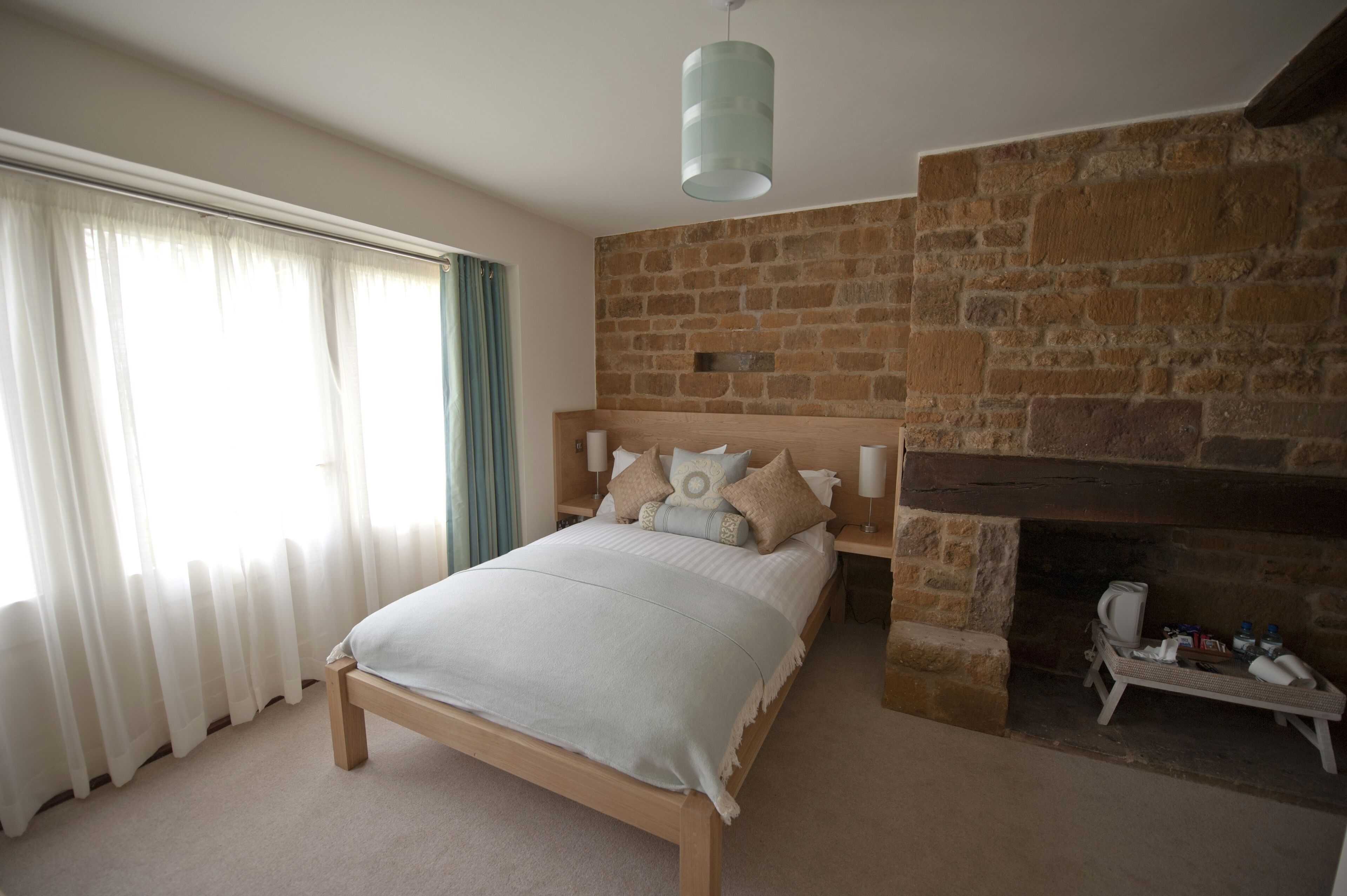 A bedroom with a stone fireplace, a bed with a wooden headboard, and a large window with sheer curtains.