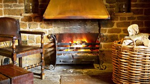 A stone fireplace with a metal grate, a wooden chair, and a wicker basket filled with logs. - The Marquess Of Exeter (Oakham)