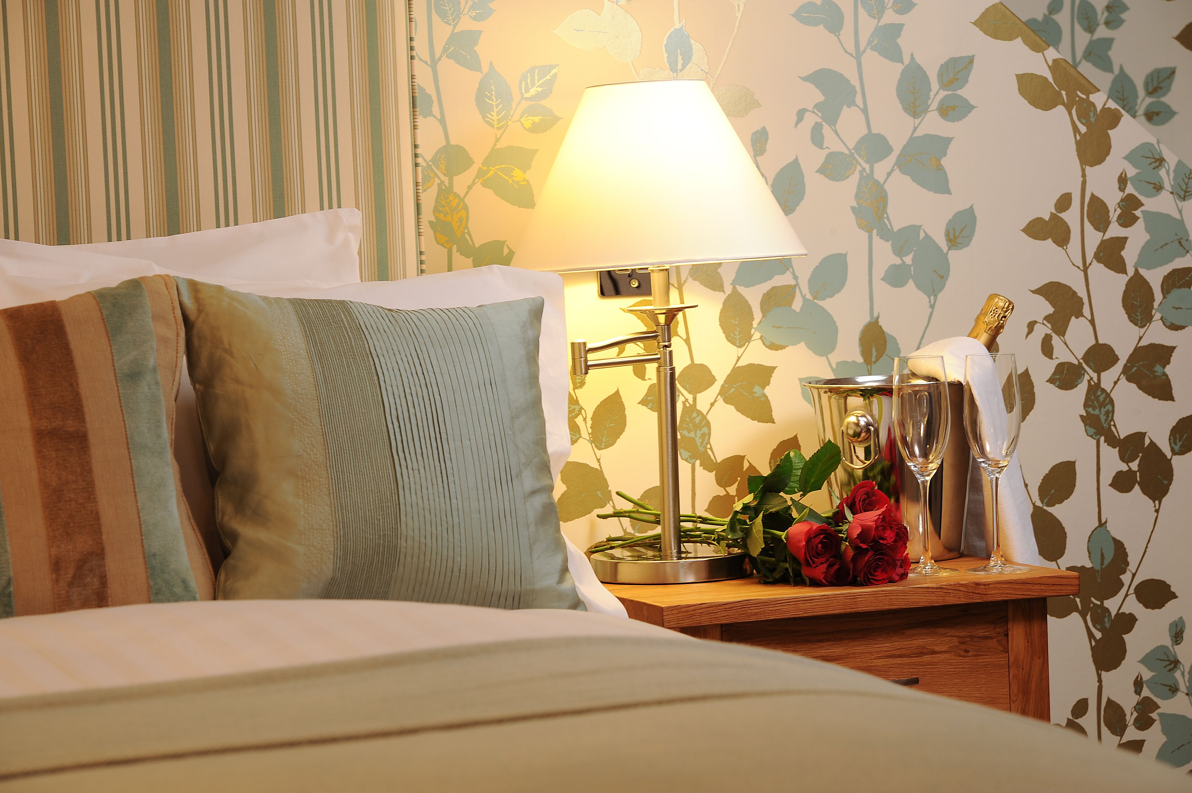 A neatly made bed with pillows, a bedside table with a lamp, a bottle, and a glass of water.