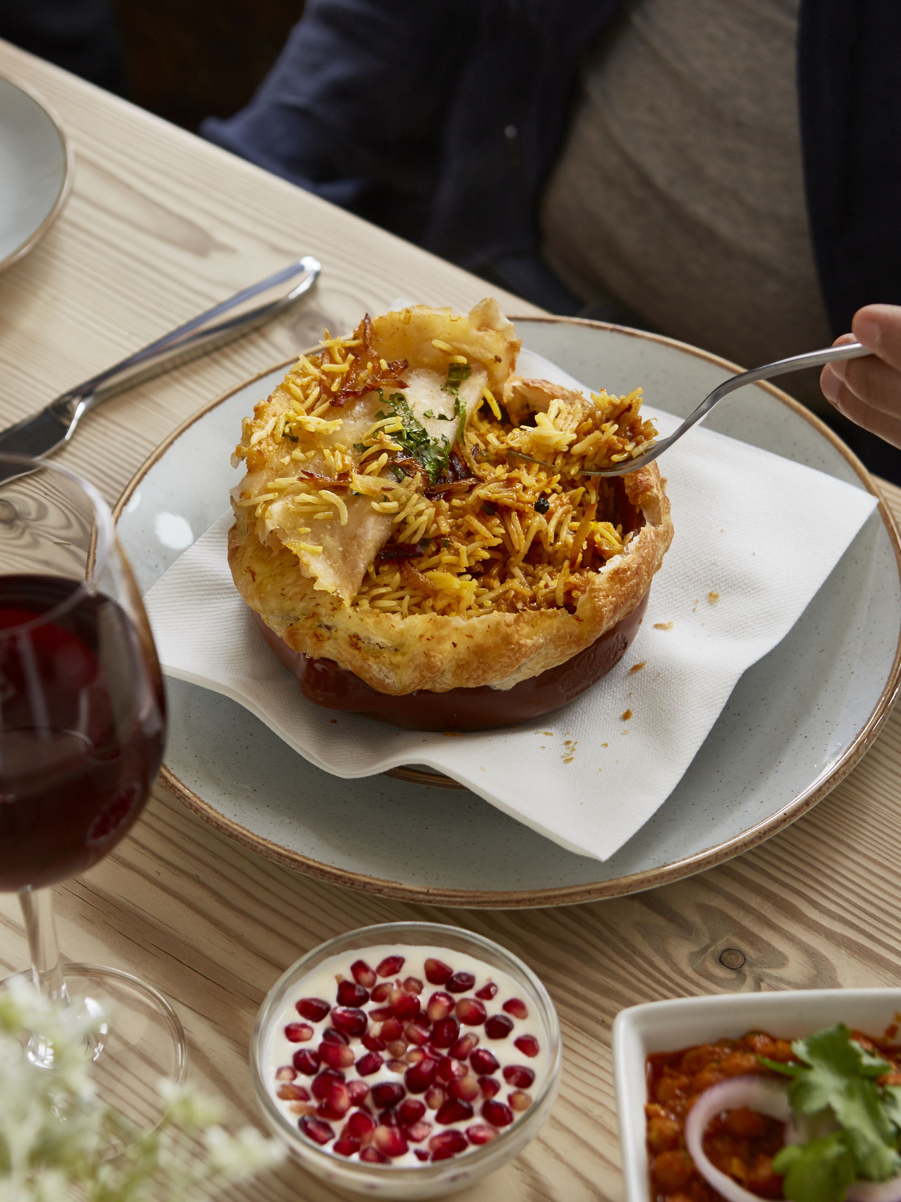 A meal with rice and meat on a plate, a glass of red wine, and a bowl of pomegranate seeds.