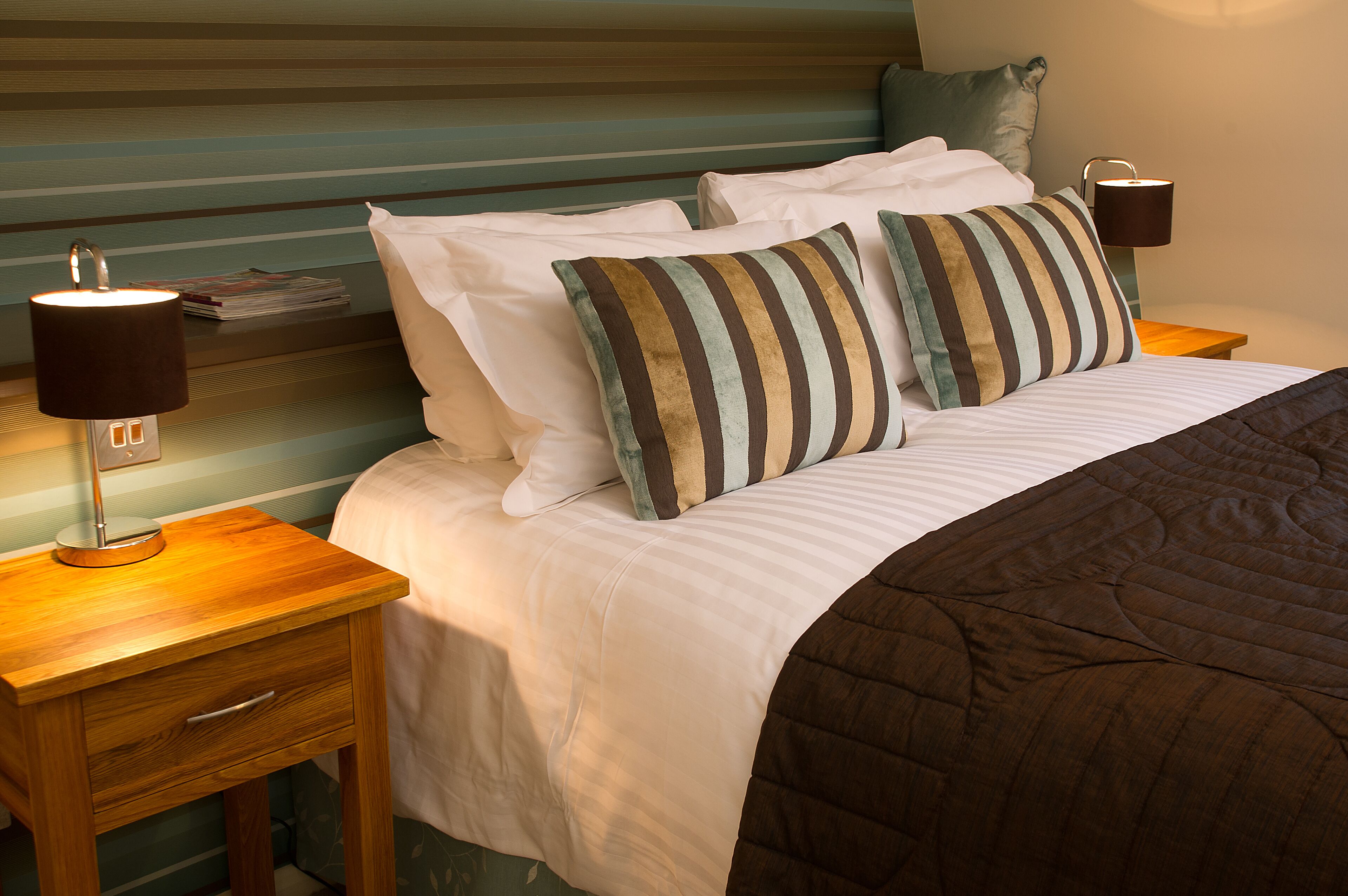 Een net opgemaakt bed met gestreepte kussens, een bruin sprei en twee bedlampen op houten nachtkastjes.