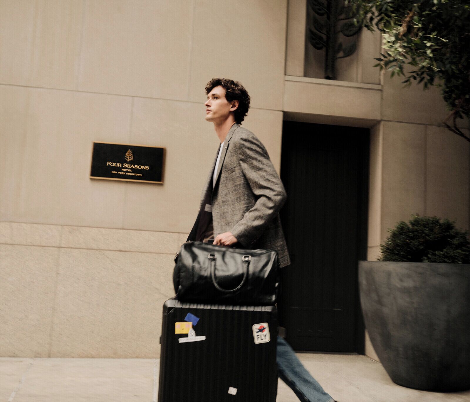 A man walking with a suitcase and a black bag, passing a sign for Four Seasons Hotel New York Downtown.