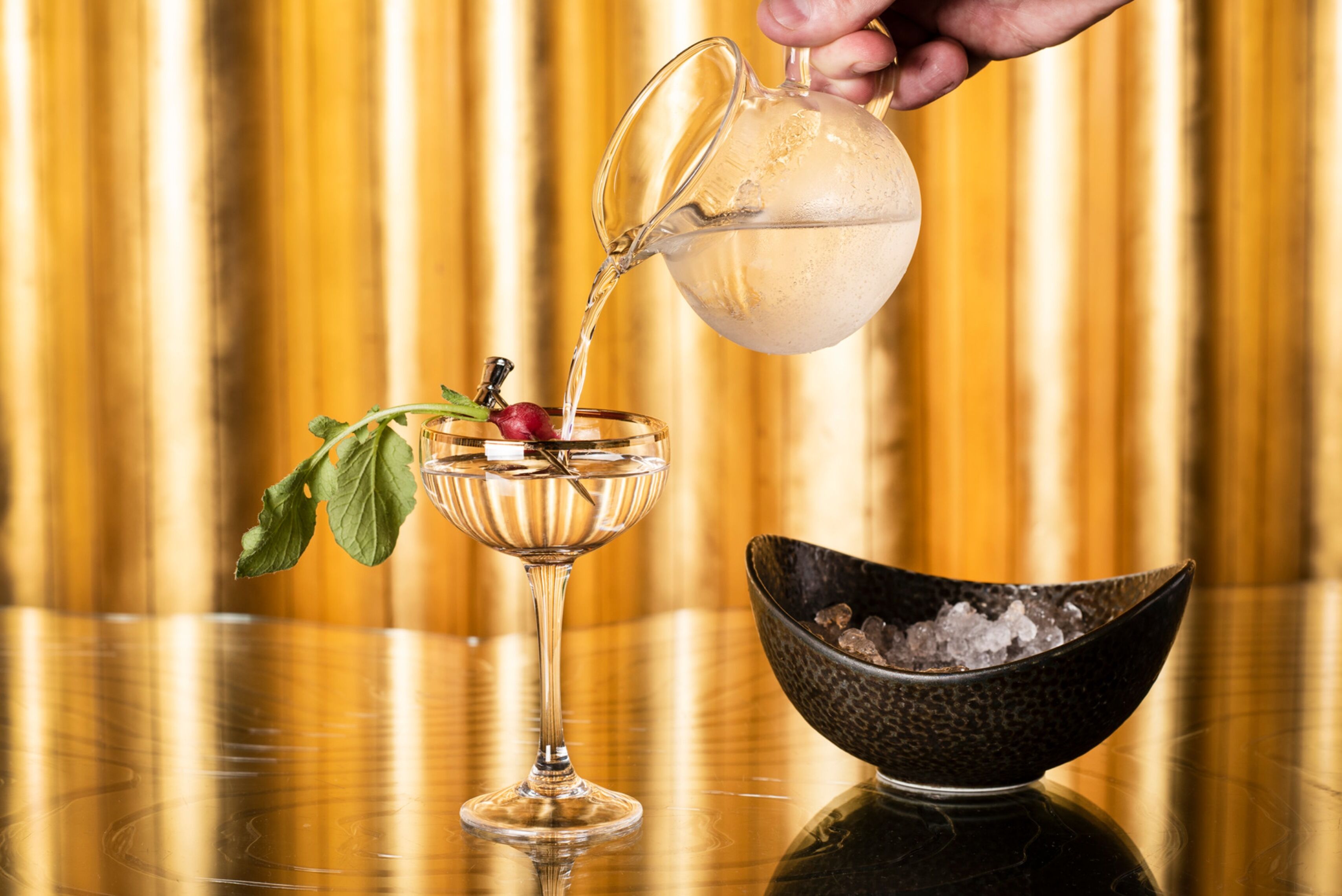 A hand pouring a clear liquid into a glass with a stemmed design, a decorative leaf, and a cherry on a stemmed glass. A bowl of ice is visible on the table.