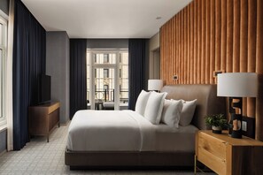 Une chambre d’hôtel avec un grand lit, une tête de lit en bois, une table de chevet avec une lampe, une télévision et une vue sur un immeuble par la fenêtre.