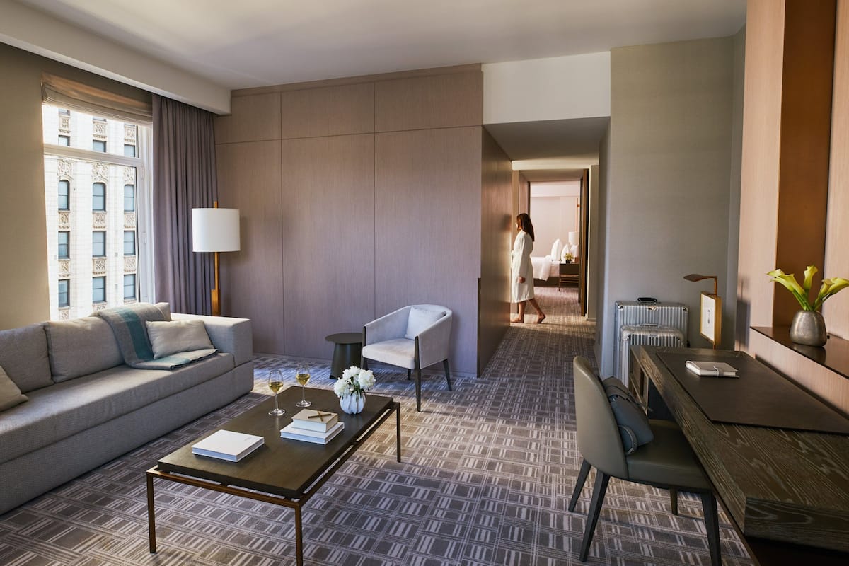 A modern hotel room with a sofa, a coffee table, a dining table, and a view of the city through a large window.