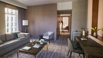 A modern hotel room with a sofa, a coffee table, a dining table, and a view of the city through a large window.