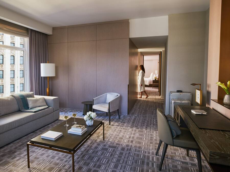 A modern hotel room with a sofa, a coffee table, a dining table, and a view of the city through a large window.