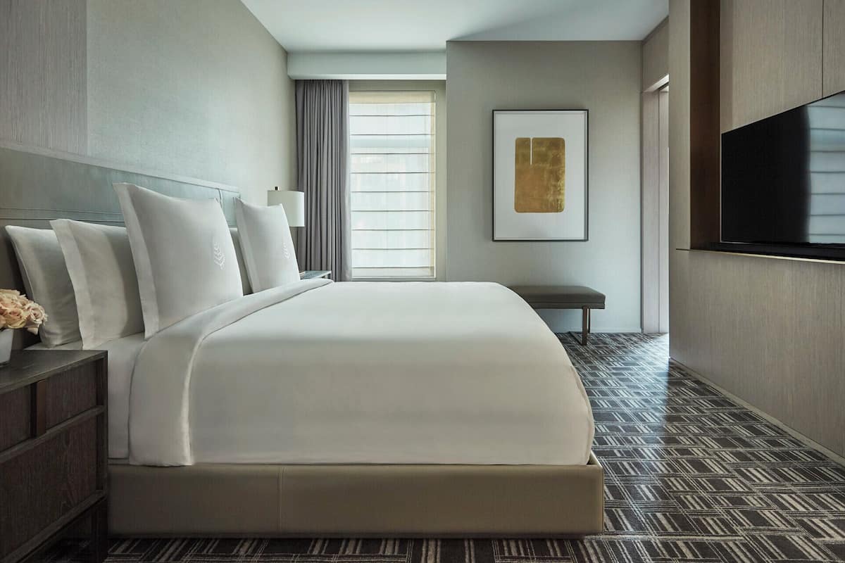 A hotel room with a large bed, a bedside table, a bench, a TV, and a window with curtains.