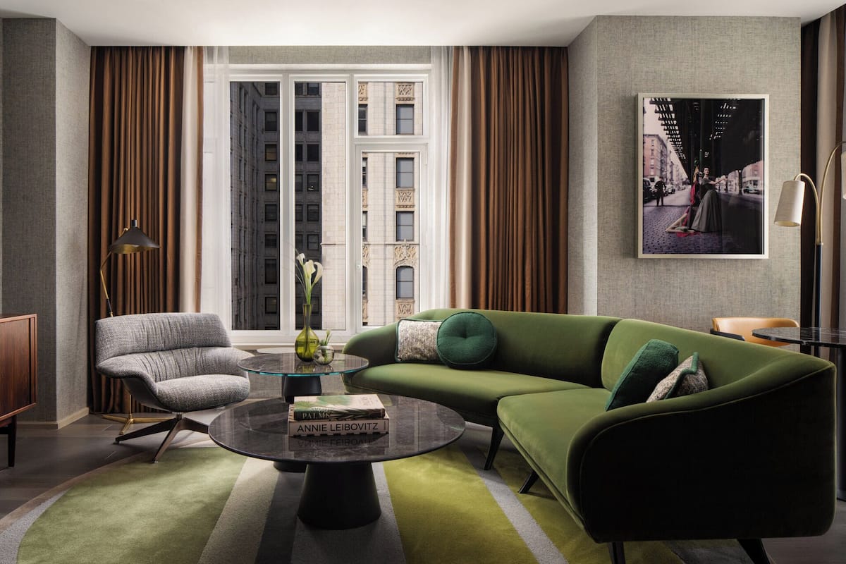 A modern living room with a green sofa, a grey armchair, and a glass coffee table. There is a large window with curtains, a painting on the wall, and a lamp on a side table.