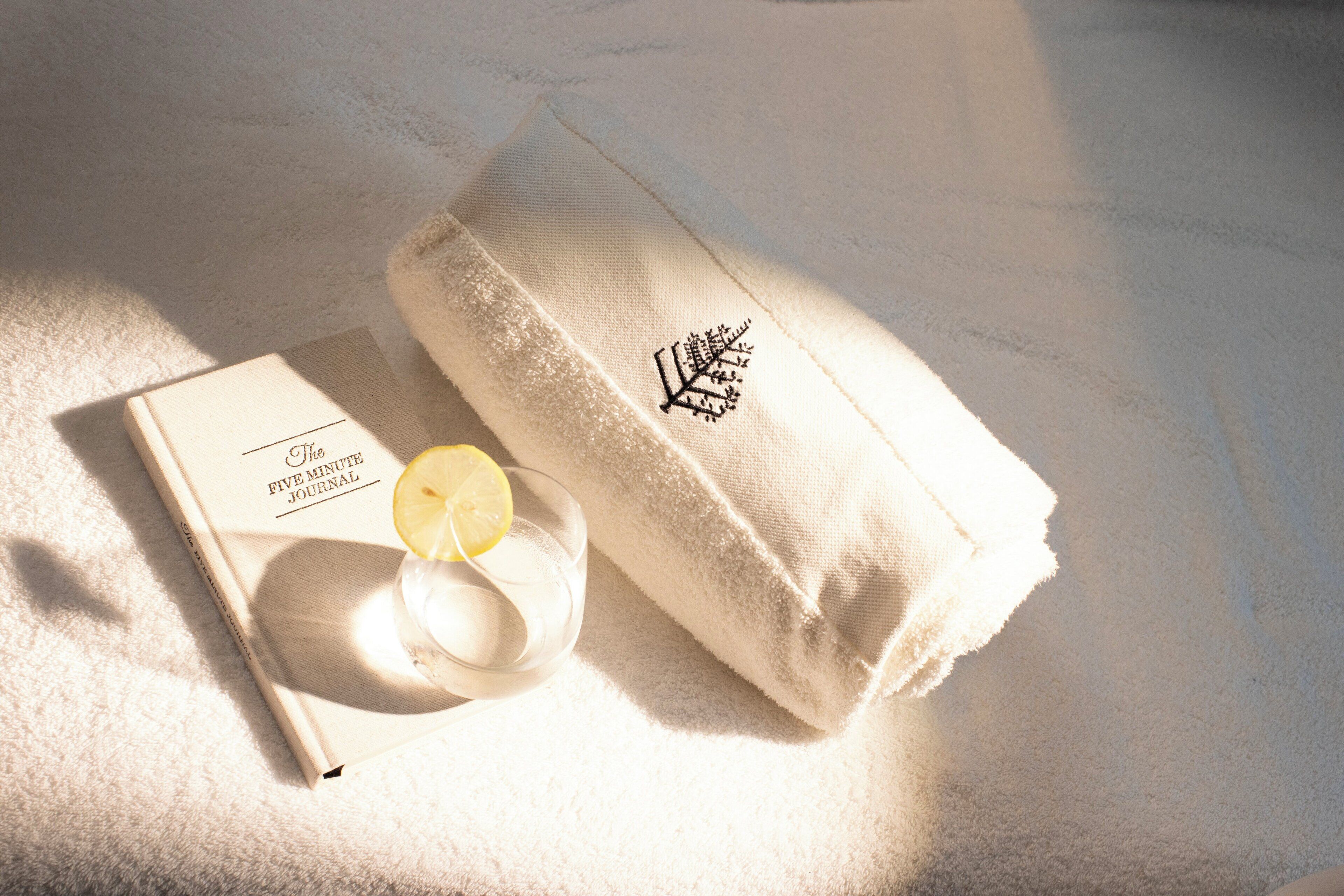 A white towel with a leaf embroidery, a glass of water with a lemon slice, and a book titled 'The Five Minute Journal' on a textured surface.