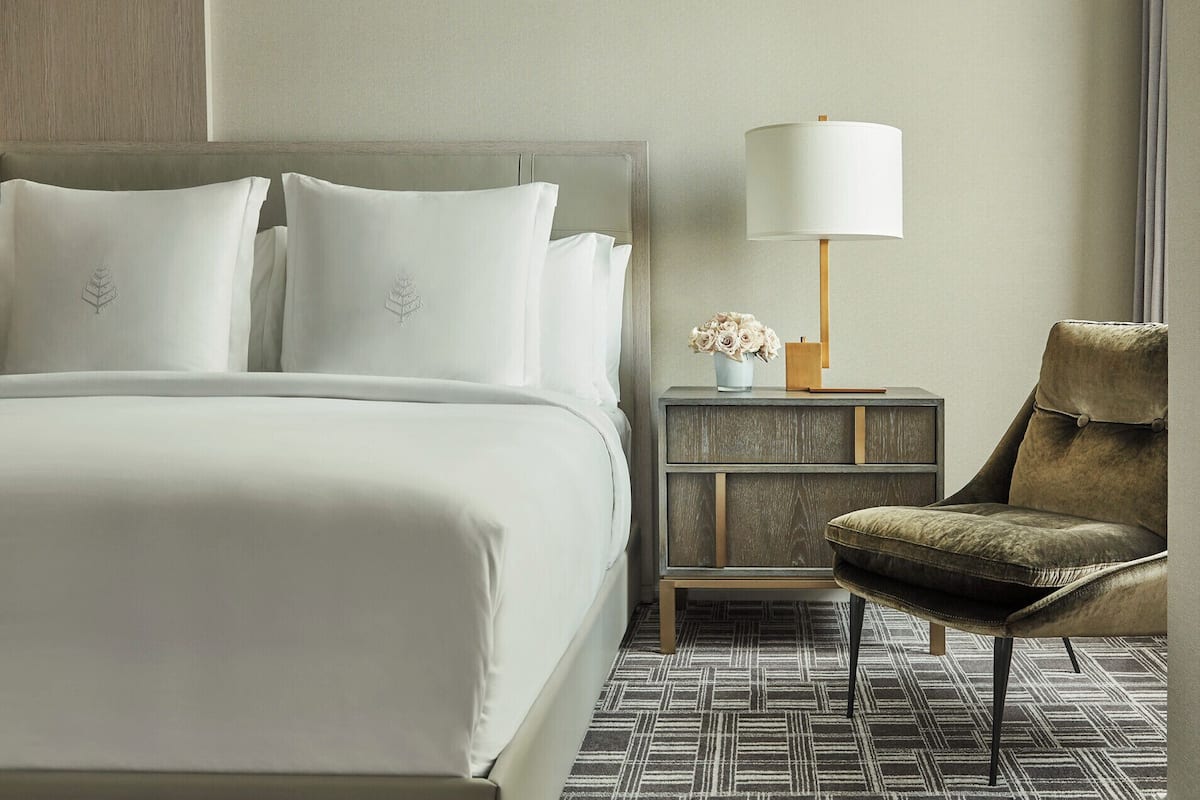 A hotel room with a bed, a nightstand with a lamp, a chair, and a patterned rug.