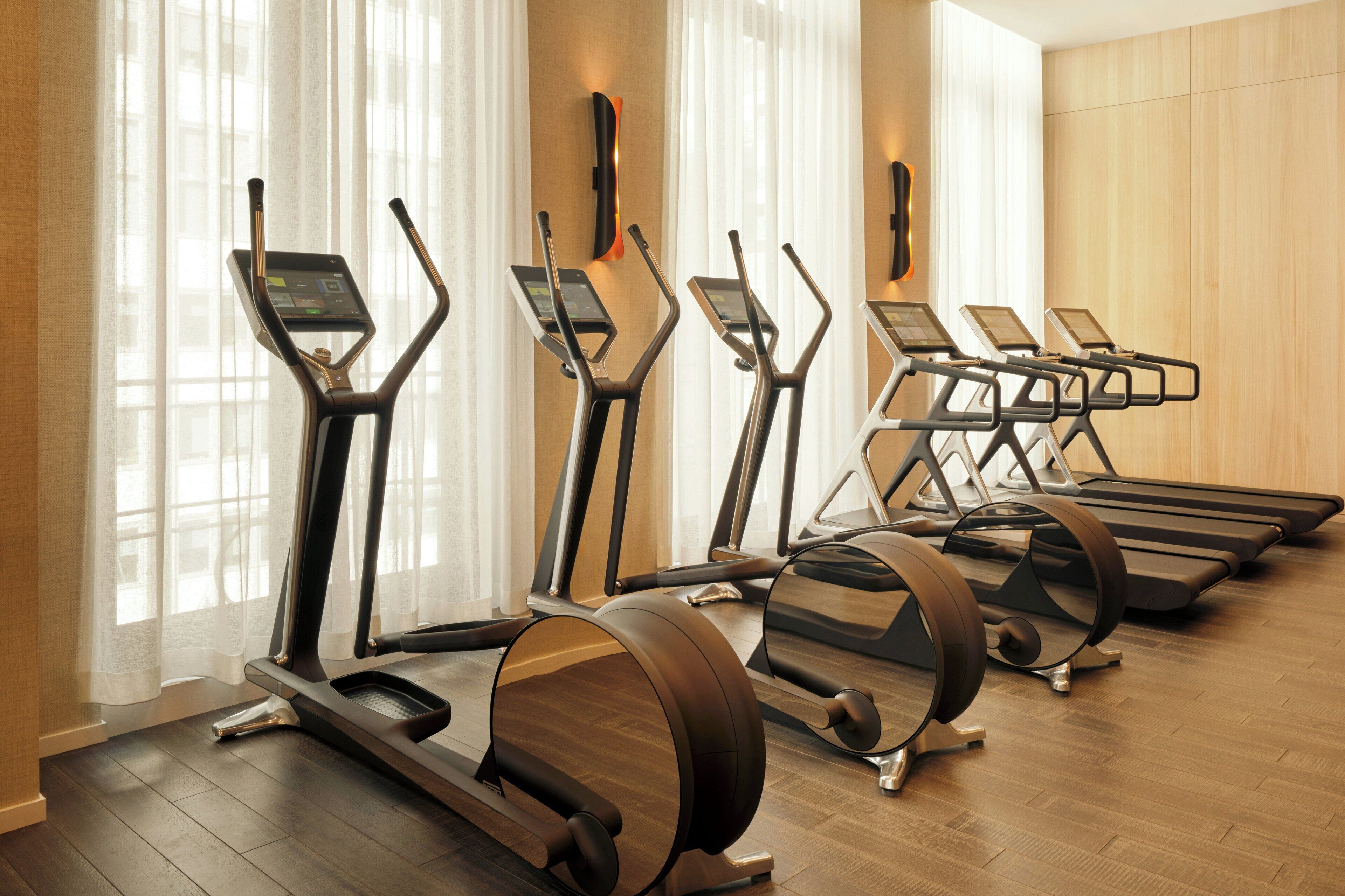 Een fitnessruimte met loopbanden en elliptische trainers, grote ramen met dunne gordijnen, en houten vloeren.