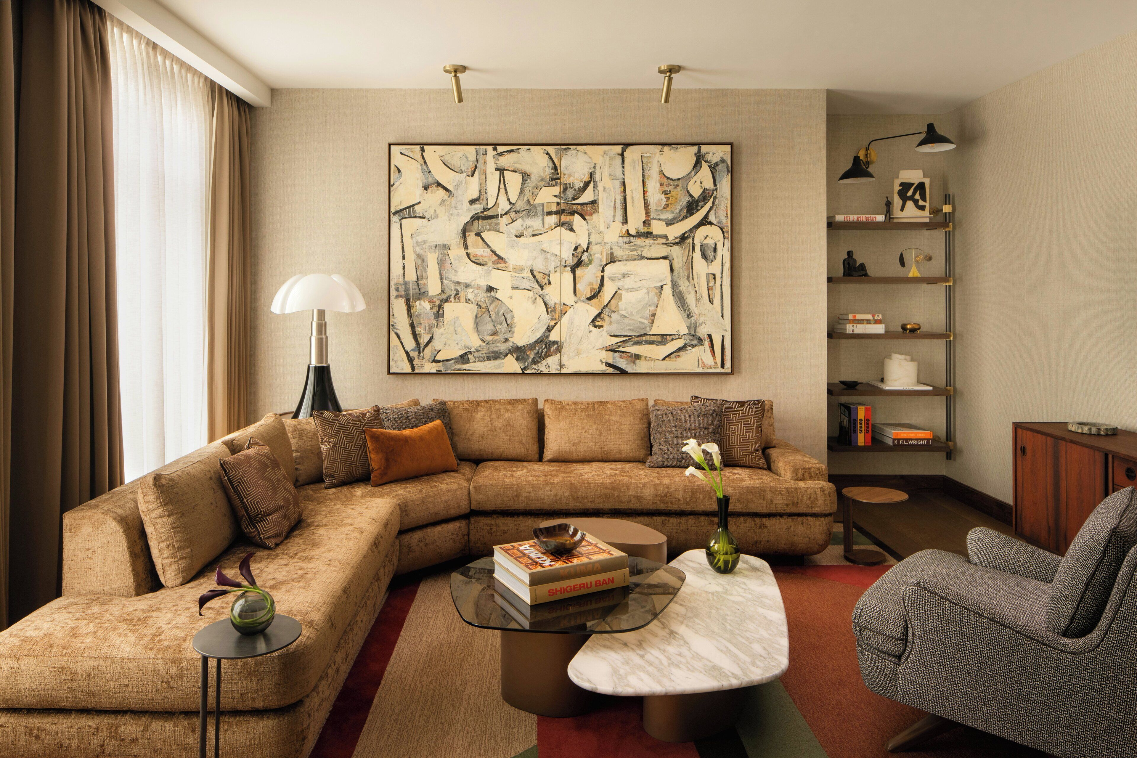 A modern living room with a sectional sofa, a glass coffee table, a side table, a reading chair, a bookshelf, and a large abstract painting on the wall.