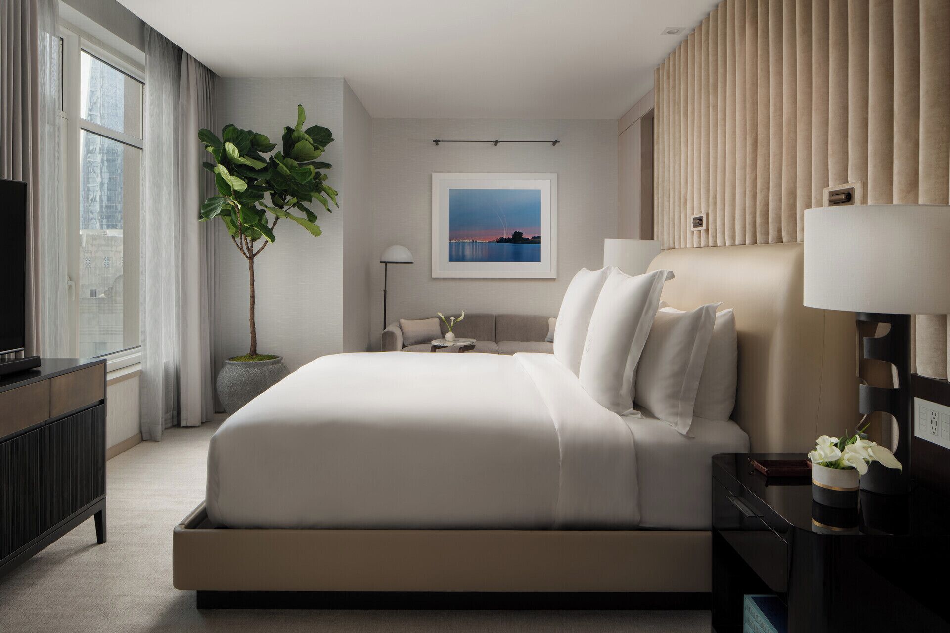 A modern hotel room with a large bed, a television, a potted plant, a framed picture on the wall, and a bedside table with a lamp.