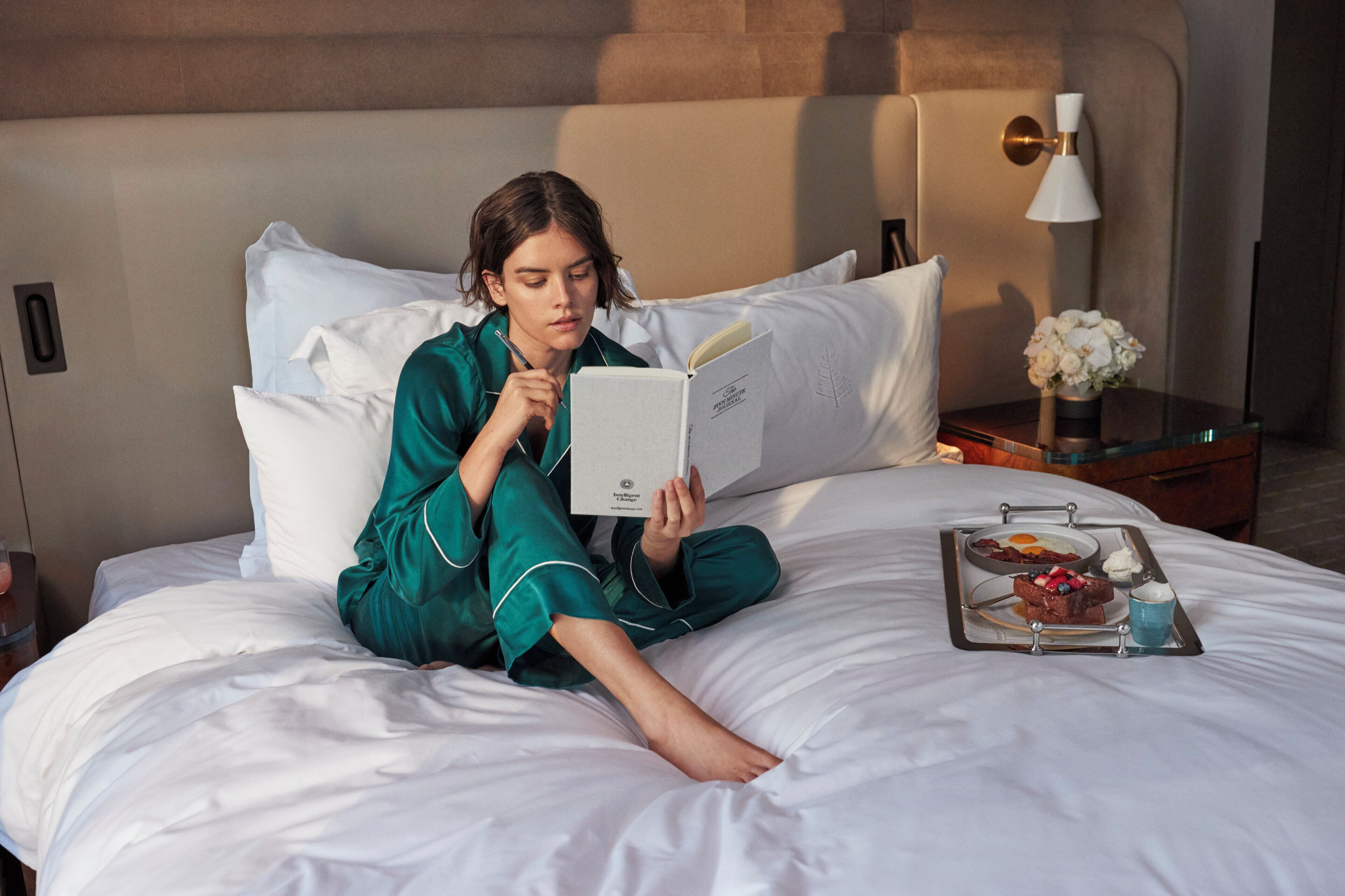 Uma pessoa sentada na cama de um quarto de hotel, lendo um livro. Há uma bandeja com comida e bebida sobre a cama.