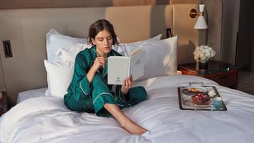 Uma pessoa sentada na cama de um quarto de hotel, lendo um livro. Há uma bandeja com comida e bebida sobre a cama.