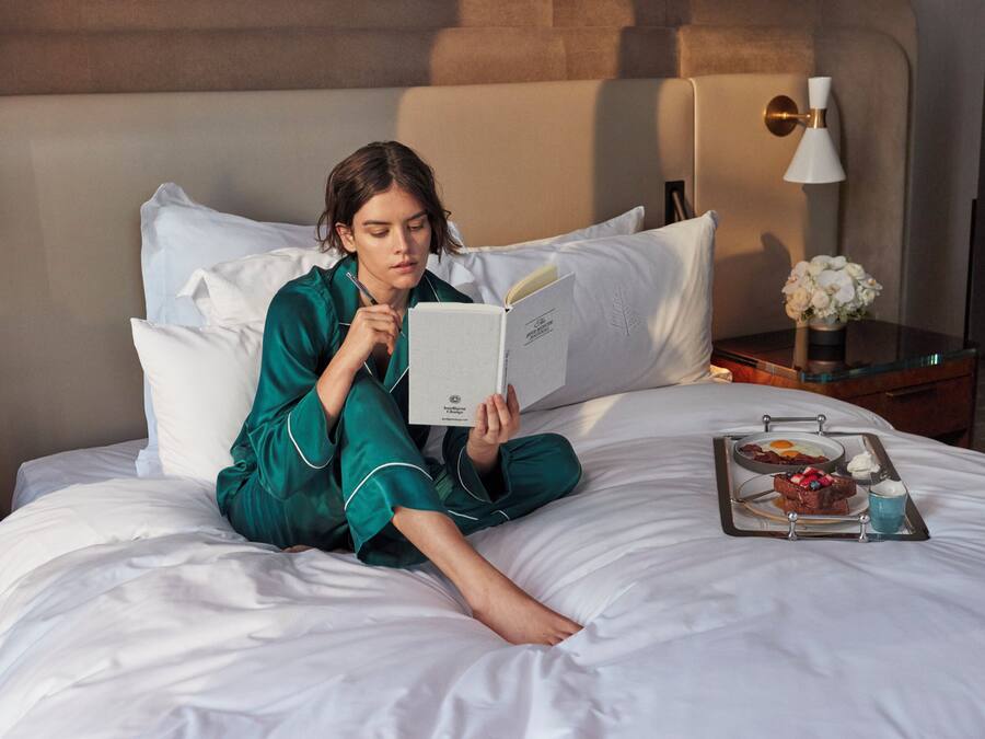 Uma pessoa sentada na cama de um quarto de hotel, lendo um livro. Há uma bandeja com comida e bebida sobre a cama.