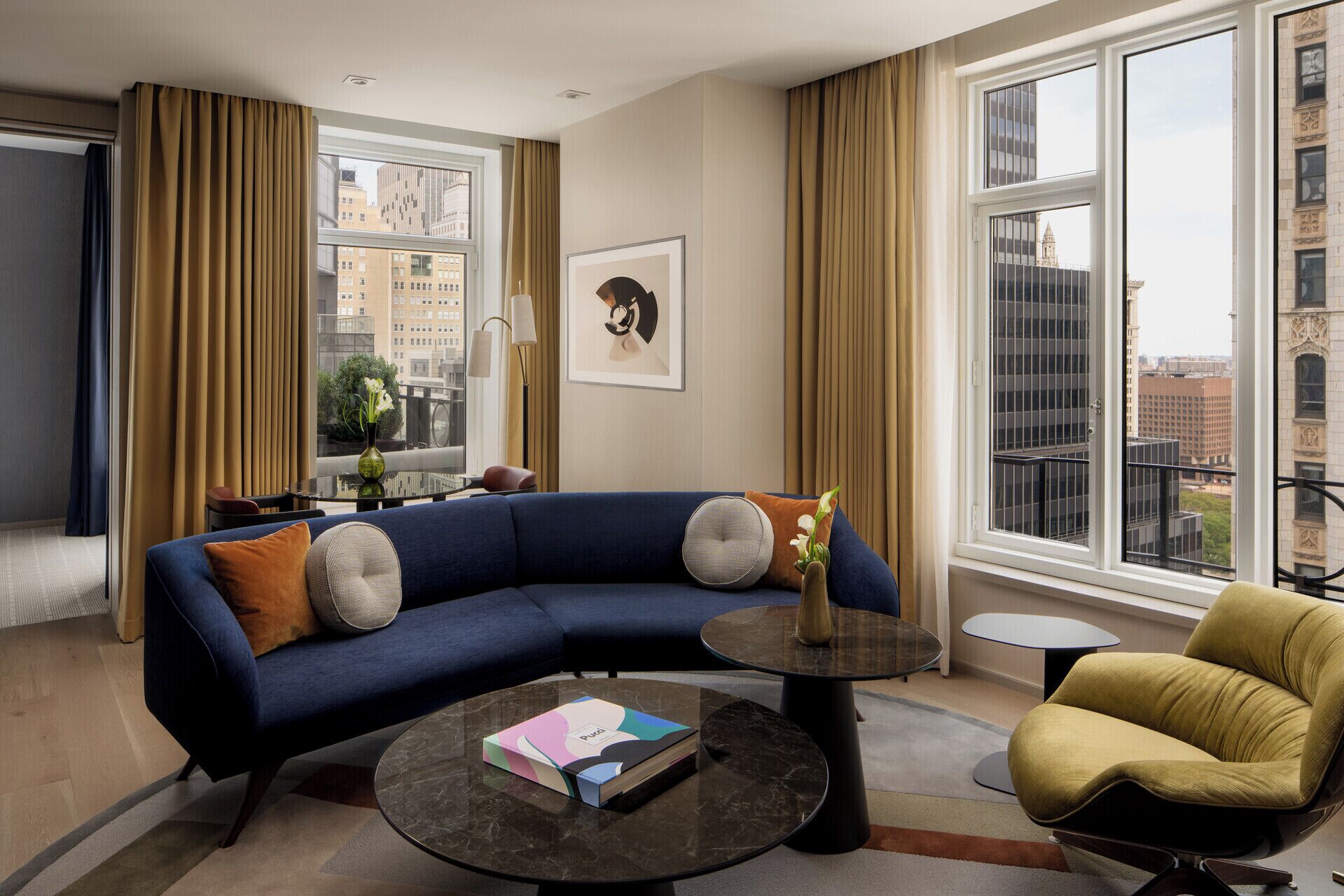 A modern living room with a blue sofa, a round coffee table, a yellow armchair, and large windows with city views.