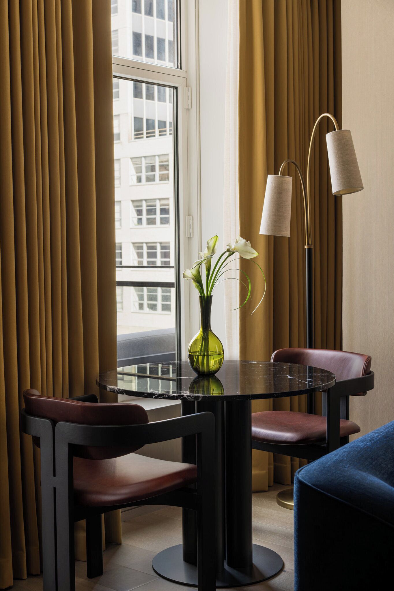 A hotel room with a dining table, chairs, a vase with flowers, a floor lamp, and a view of buildings through a window.