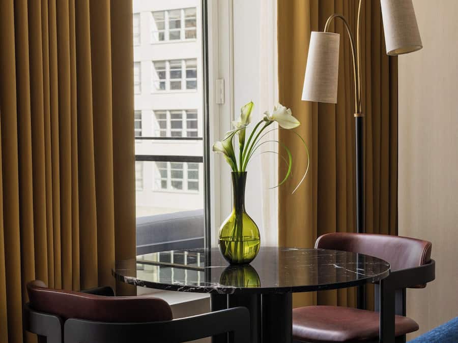 A hotel room with a dining table, chairs, a vase with flowers, a floor lamp, and a view of buildings through a window.