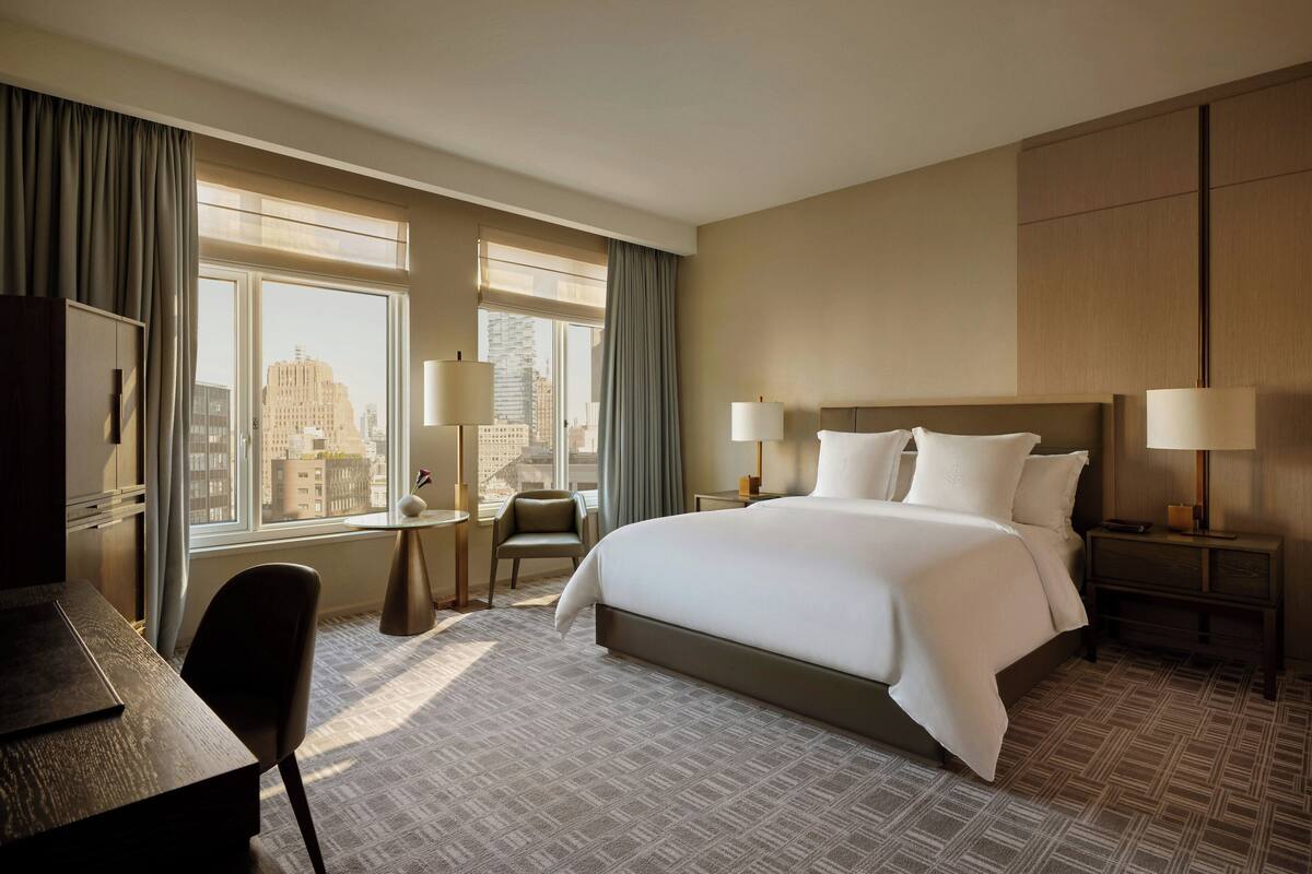 A hotel room with a large bed, two bedside tables with lamps, a desk, a chair, and a view of the city through large windows.