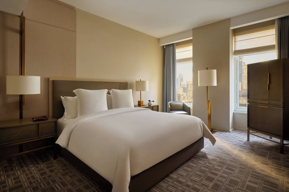 A hotel room with a large bed, two bedside tables with lamps, a wardrobe, and a view of the city through the window.