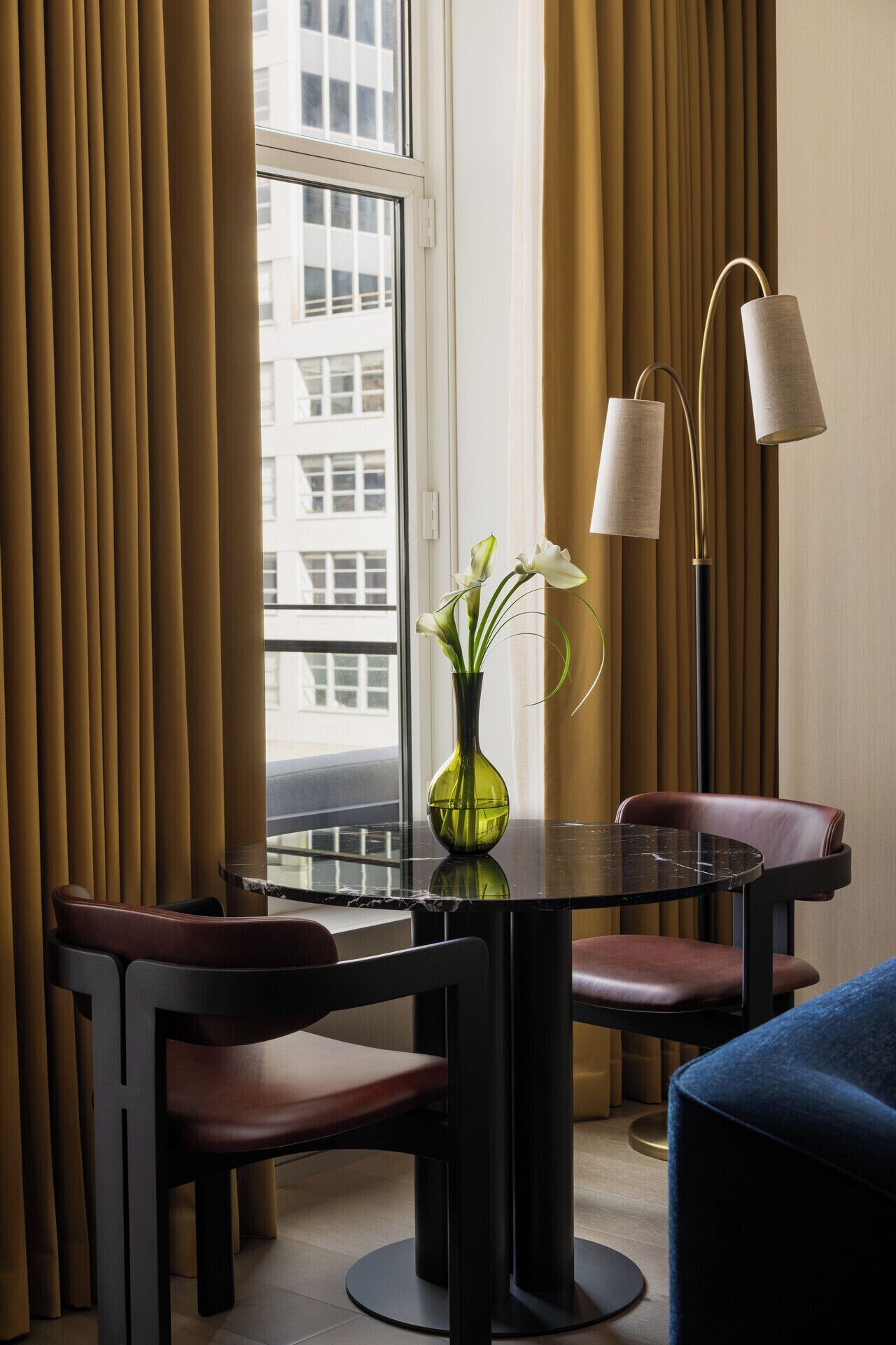 Een hotelkamer met een eettafel, stoelen, een vaas met bloemen, een vloerlamp en uitzicht op de stad door een raam.