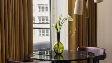 Een hotelkamer met een eettafel, stoelen, een vaas met bloemen, een vloerlamp en uitzicht op de stad door een raam.