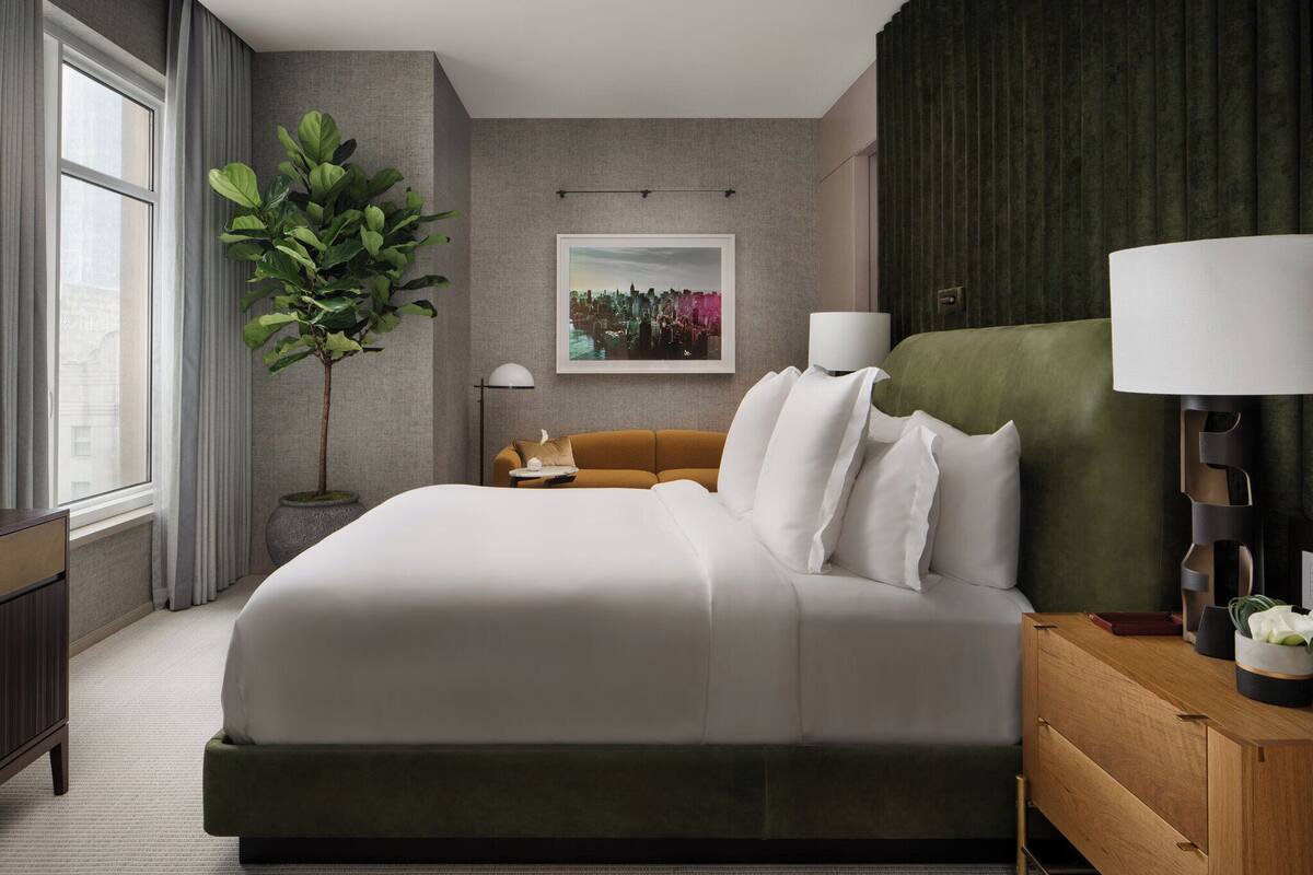 A hotel room with a large bed, a green headboard, a side table with a lamp, a potted plant, and a framed cityscape picture on the wall.