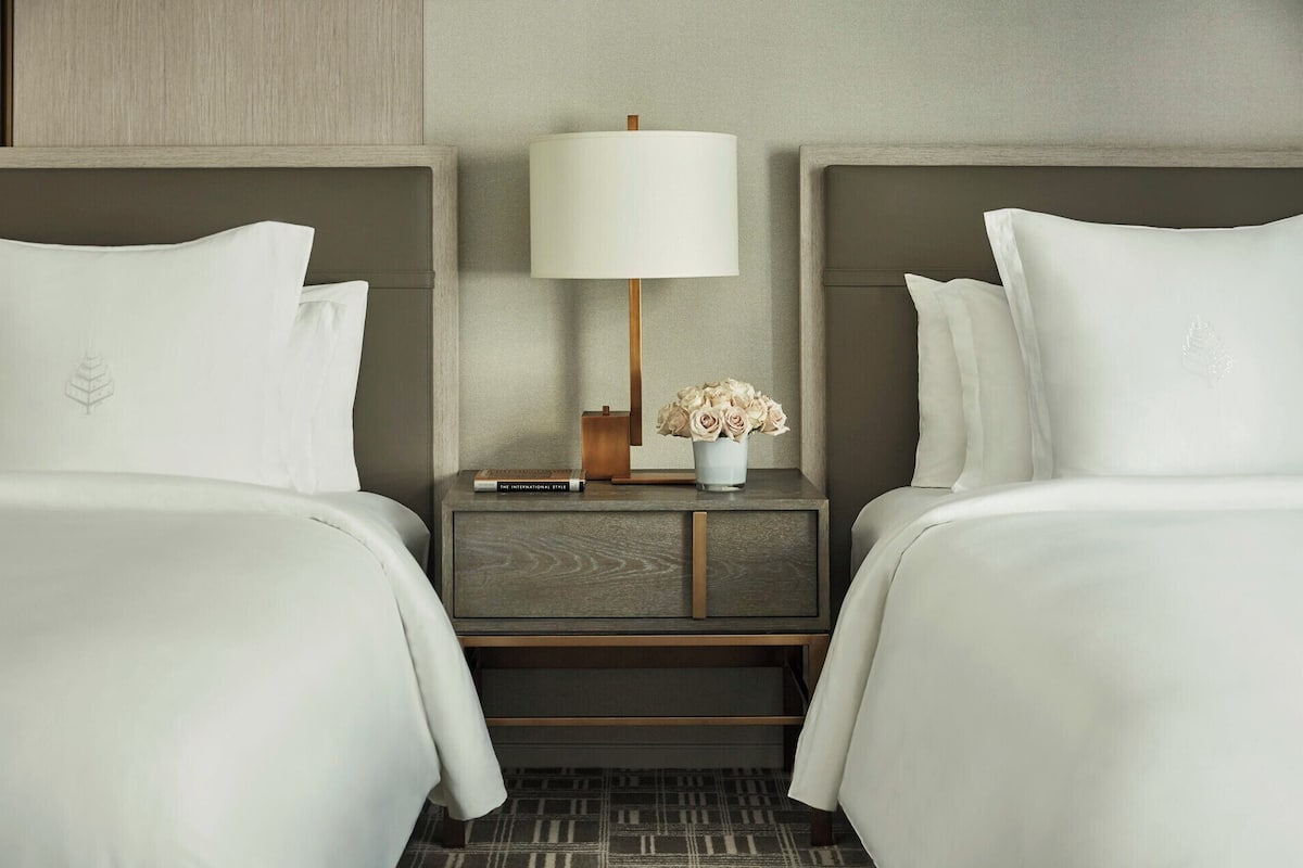 A hotel room with two beds, a nightstand with a lamp, a vase with flowers, and a book titled 'The International Style'.