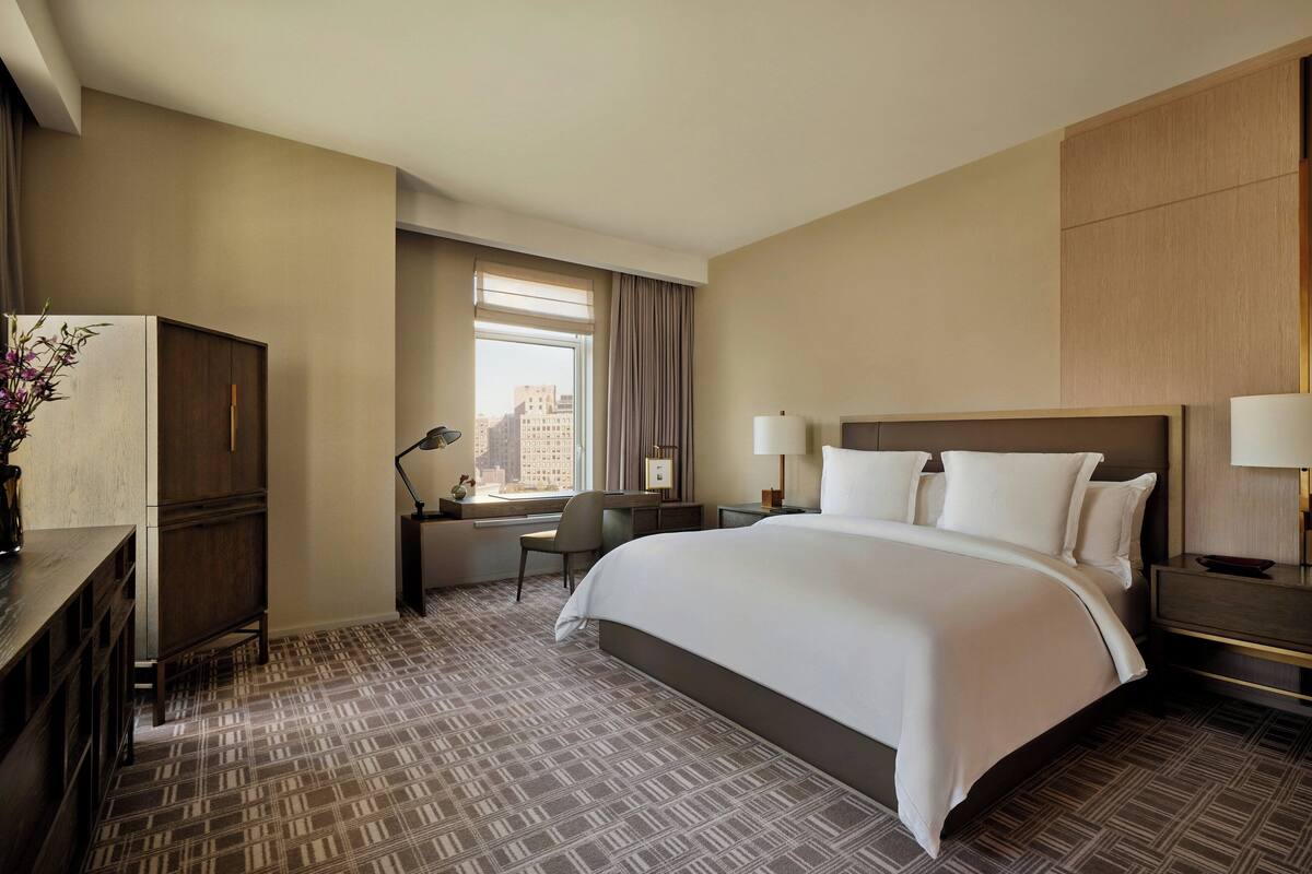 A hotel room with a large bed, a desk with a chair, a desk lamp, a nightstand with a lamp, and a view of the city through a window.