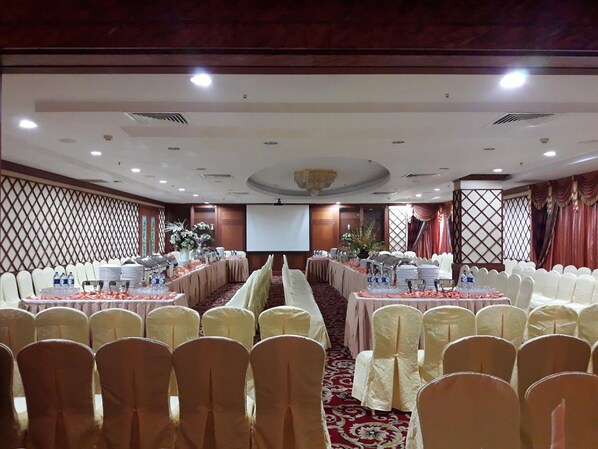 Un salón de banquetes con filas de sillas blancas, mesas con botellas de agua y vasos, y una pantalla de proyector.